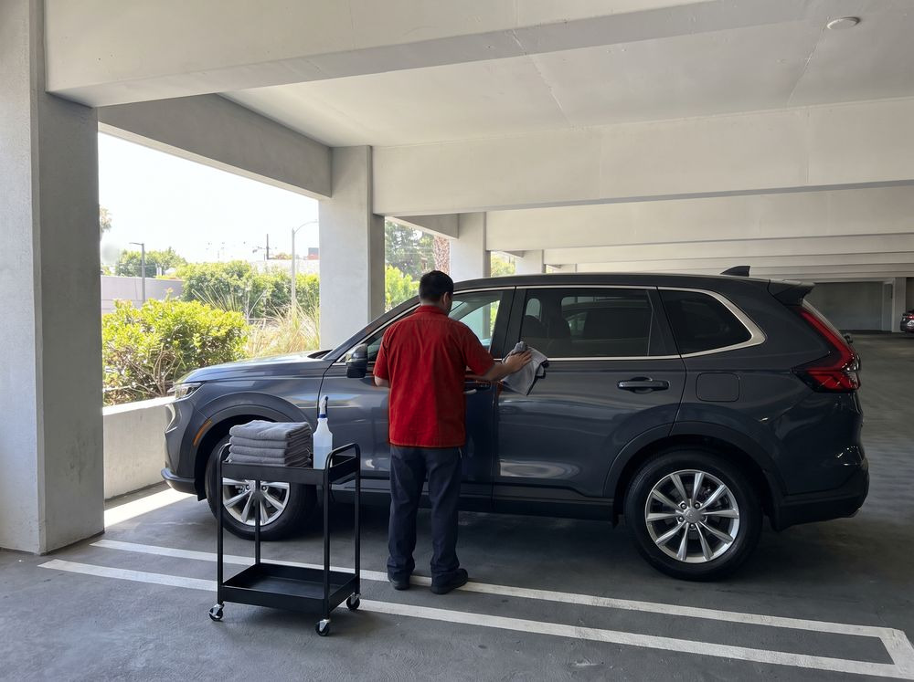 A technician in a red shirt wiping down a gray SUV in an apartment building parking garage, beside a cart of folded towels